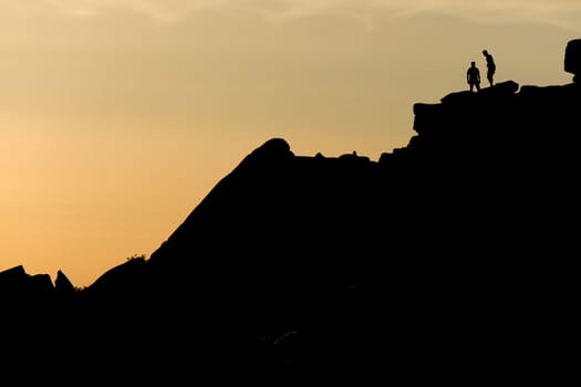A pair of rock climbers, silhouetted at sunset, look back from the summit of stange edge. Located in the peak district in england. Outdoor activity