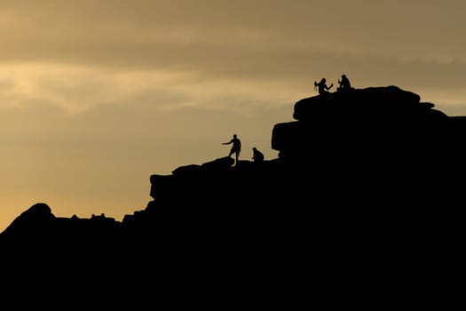 People on stanage edge, peak district, at sunset. A couple take a selfie holding drinks, silhouetted against the golden sky. Climbers stand there too