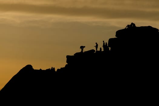 Crowds of people, including a romantic couple, surround a climber at the top of stanage edge in the peak district. Tourists enjoy the outdoors