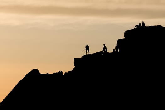 Dramatic silhouette scene as a climber sorts his ropes at the top of a striking rock formation. Other people enjoy the sunset and remote wilderness.