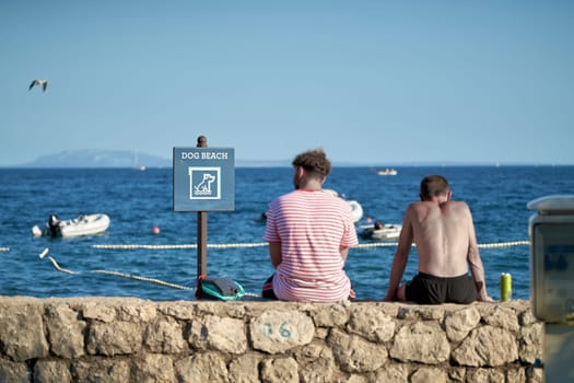 Sign with the inscription dog beach on a campsite in Krk in Croatia. Next to it two vacationers resting on a wall.