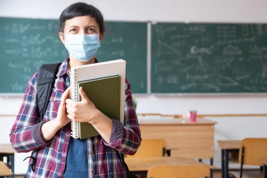 Smiling student girl wearing school backpack, mask and holding exercise book. Social distanting and classroom safety during coronavirus epidemic