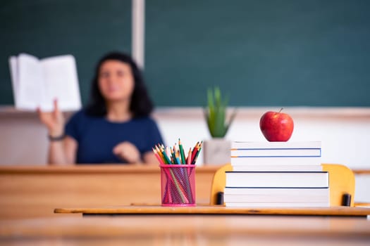 Back to School Concept. Apple, accessories and books in the classroom, chalkboard and teacher teaching in the background.