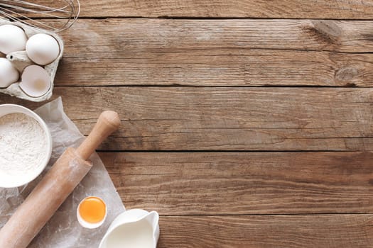 Baking ingredients, kitchen utensils on old wooden background. Cooking dough, preparing egg yolk, flour, rolling pin, milk, parchment paper, whisk, salt, sodium. Concept flat lay photo with copy space