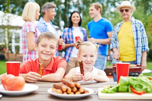 Cute siblings with sausages eating outdoors with their parents on background
