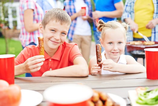Brother and sister eating sausages outdoors with parents on background