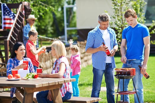 Two men frying sausages on grill outdoors with young women talking by table near by
