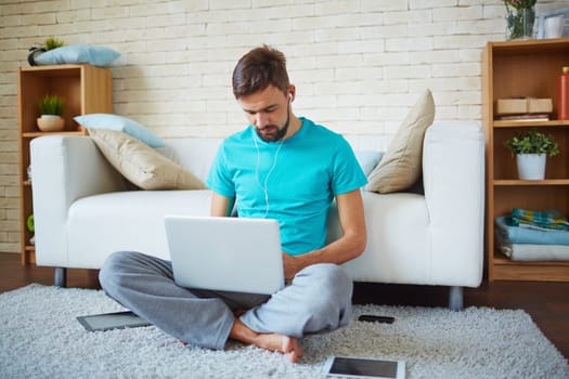 Young man listening to music in earphones while networking on laptop at leisure