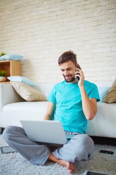Happy man listening to music in earphones, speaking on mobile phone and networking on laptop