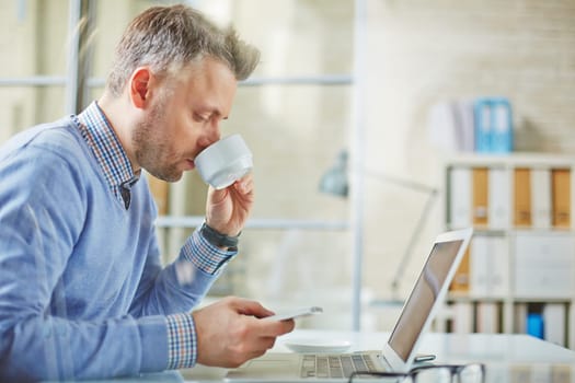 Businessman drinking tea while reading sms on cellphone before meeting with partners