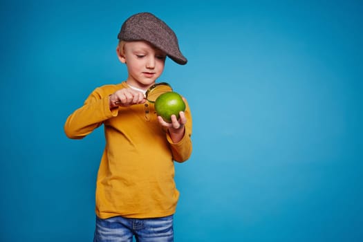 Cute schoolboy in casualwear and cap looking at green apple through magnifying glass