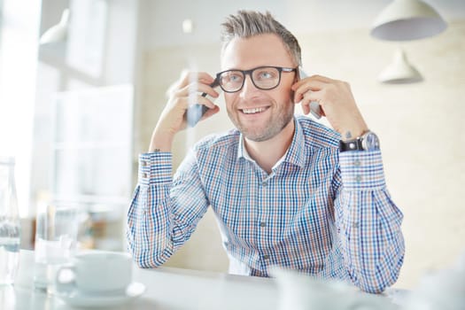 Smiling businessman in eyeglasses holding two smartphones by ears