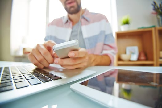 Hands of young man using smartphone over laptop keypad