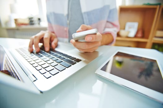 Hands of young man holding smartphone and typing on laptop
