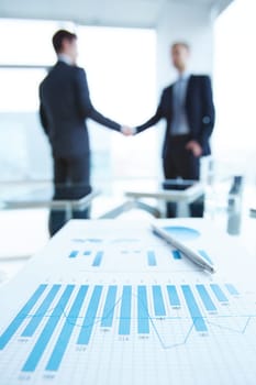 Business document with chart and graph on workplace with two men handshaking on background