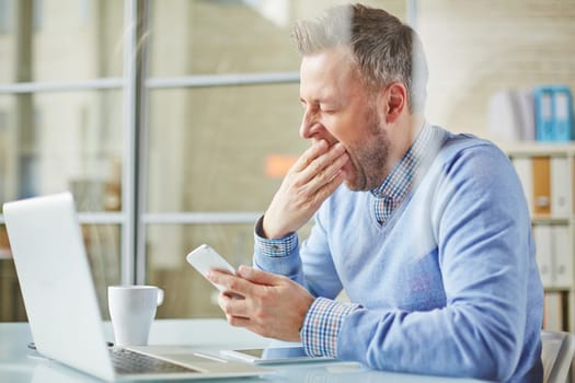 Tired businessman with cellphone yawning at workplace