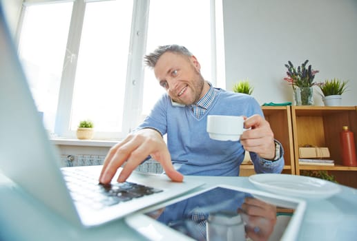 Businessman with coffee working on laptop and speaking on cellphone in office