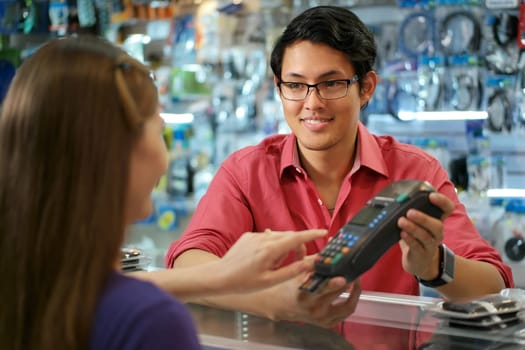 Young woman paying with credit card in computer shop, with asian sales clerk smiling and holding pos. The girl enters security code and waits for receipt