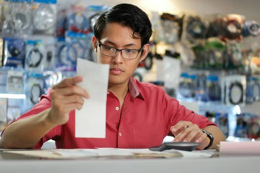 Young asian entrepreneur owning a computer shop and checking bills and invoice with calculator, doing budget and reviewing expenses.