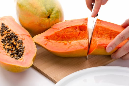 First cut through a hollowed out half of a papaya. Finger tips of the left hand of a chef are fixing the fruit on a wooden cutting board, while his right is guiding a kitchen knife. Cutout. Close up.