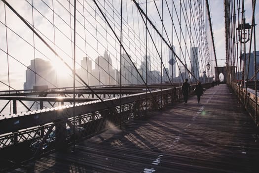 Couple walking on pedestrian path across Brooklyn bridge. New York City Manhattan downtown skyline in sunset with skyscrapers illuminated over East River panorama as seen from Brooklyn bridge.