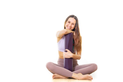 Young sporty female yoga instructor in bright white yoga studio, holding yoga mat, smiling, looking at camera.