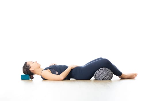 Restorative yoga with a bolster. Young sporty attractive woman in bright white yoga studio, lying on bolster cushion, stretching and relaxing during restorative yoga. Healthy active lifestyle.