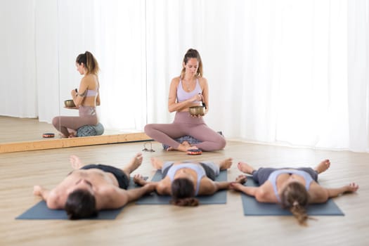 Yoga instructor and group of young sporty people in yoga studio, lying and relaxing on yoga mats during restorative yoga session. Healthy active lifestyle.