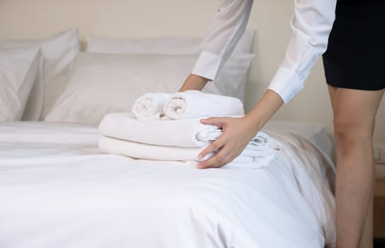 Hotel room service making room. Woman chambermaid keeping stack of fresh white bath towels on bed in guest room
