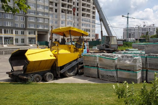 An asphalt paver is parked at a construction site. Road construction machinery stands by the road in a warehouse. Construction of heavy roads and repair of city streets