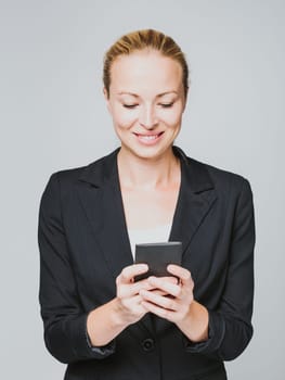 Beautiful young caucasian businesswoman in business attire using smart phone application. Studio portrait shot on gray background.