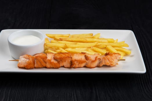 Grilled salmon fish shashlik with fried potatoes and sauce on white plate on black wooden background