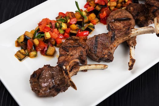 Roasted lamb ribs with vegetables on white plate on black wooden background