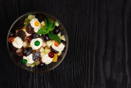 Salad from grapes apples pears kiwi oranges with mascarpone chease and cream. Healthy fresh fruit summer salad in glass bowl on black wooden background with copy space.