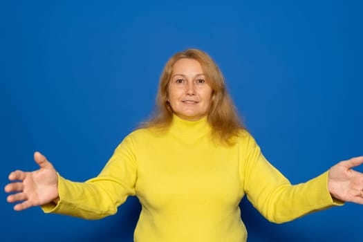 Caucasian woman in her 40s wearing a yellow sweater with her arms open in a welcoming gesture, isolated on blue studio background