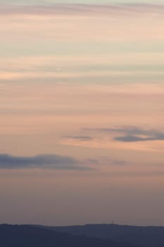 Vertical hazy sunset. Horizon under warm and faded pastel colours. Gently clouded sky above the forested horizon with protruding telecom towers.