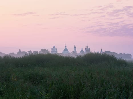 Lilac-pink sky at dawn before sunrise. A distant view of the domes of an Eastern Orthodox monastery in the morning fog behind a grassy hill. Russia, the Rostov's outskirts from the side of the Monastery of St. Jacob Saviour (Spaso-Yakovlevsky).
