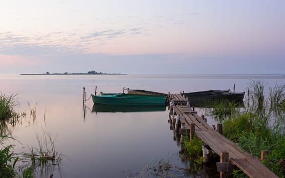 Beautiful lilac-pink dawn over a calm lake. Rowing boats are moored to a rural wooden pier at the grassy shore. Image of a serene morning landscape with space for copy. Russia, Yaroslavl Oblast, Lake Nero.