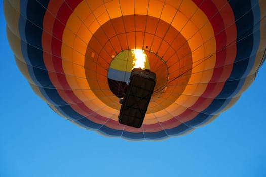 view from the ground of colorful hot air balloon with large wicker passenger basket rising up in the blue sky using burner fire flame