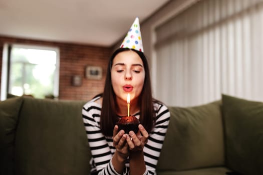 Birthday girl brunette in cardboard hat sits on the couch or sofa and blows out burning candle on cupcake. Woman holding birthday cake in her hands. Congratulation concept with blurred background