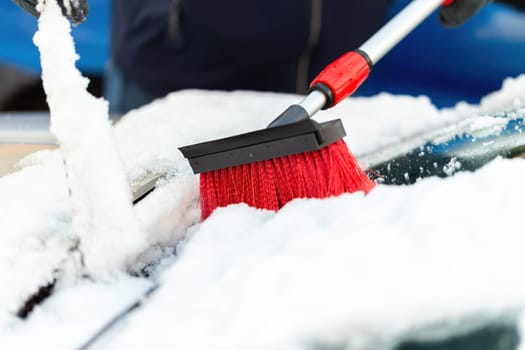Person with broom brushing snow from windshield in close-up. Hand with cleaning eqipment removing snowflakes from car. Cleaning white frost from automobile.