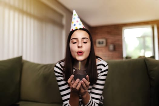 Birthday girl brunette in cardboard hat sits on the couch or sofa and blows out burning candle on cupcake. Woman holding birthday cake in her hands. Congratulation concept with blurred background