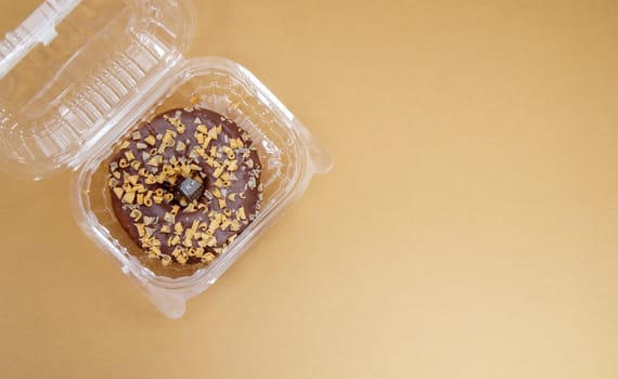 Chocolate donut in a plastic container on a brown or coffee background. Takeaway breakfast concept. One donut is packed in a plastic box for delivery. Sweet pastries delivered to your home. Top view