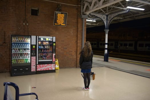 Doncaster, United Kingdom, 22nd May, 2021: Singular young woman wearing a coat, skirt and hand bag, waits during night time at doncaster train station