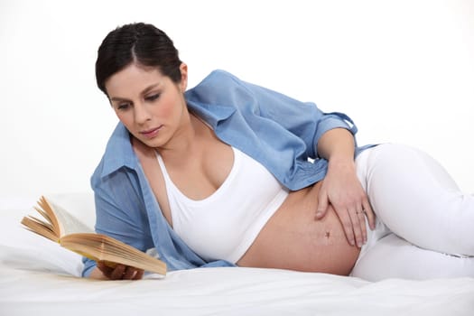 A pregnant woman reading a book on her bed.