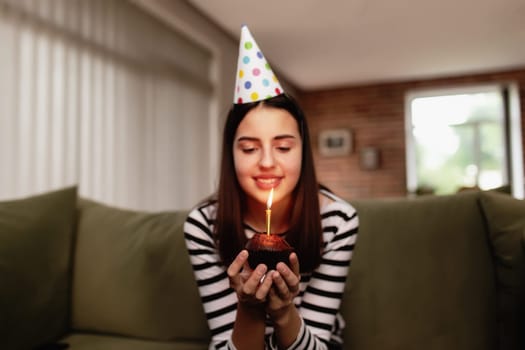 Birthday girl brunette in cardboard hat sits on the couch or sofa and blows out burning candle on cupcake. Woman holding birthday cake in her hands. Congratulation concept with blurred background