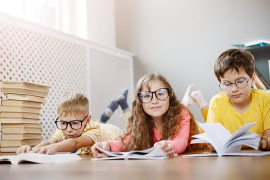Group of cute children in glasses lying on the floor indoors with books. Concept of hobby and education.