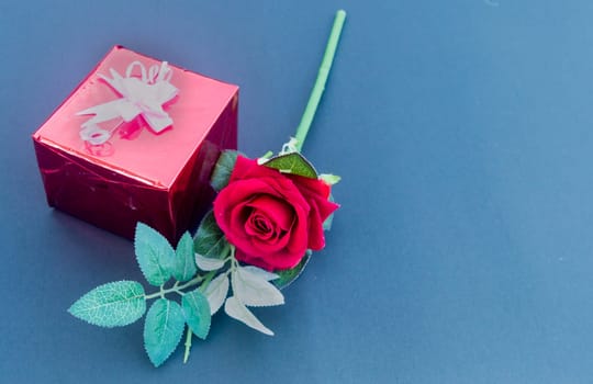 A red rose and a wrapped gift box with ribbon on table in blue background. High angle view. Flat Lay. Copy space room for text.