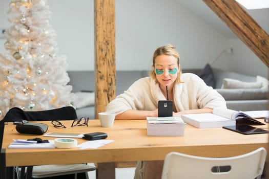 Young businesswoman wearing cosy warm bathrobe working remotly from home in winter Christmas time during 2020 corona virus pandemic. Work from home, selter in place, concept.