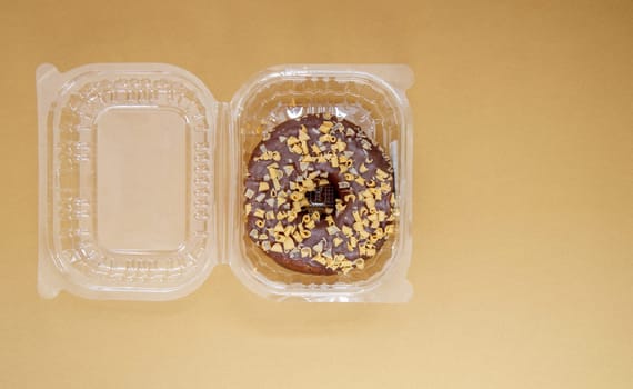 Chocolate donut in a plastic container on a brown or coffee background. Takeaway breakfast concept. One donut is packed in a plastic box for delivery. Sweet pastries delivered to your home. Top view